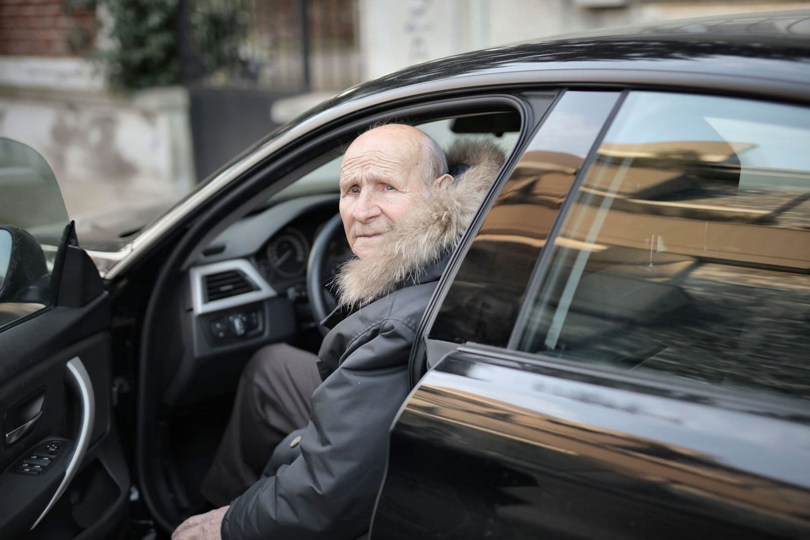 An older man seated in a car with the door open, about to step out onto the street