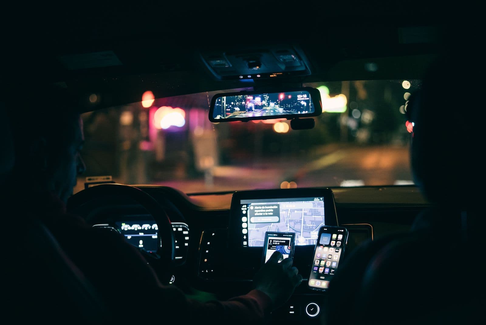 A driver at night navigating with a phone mounted beside the dashboard screen