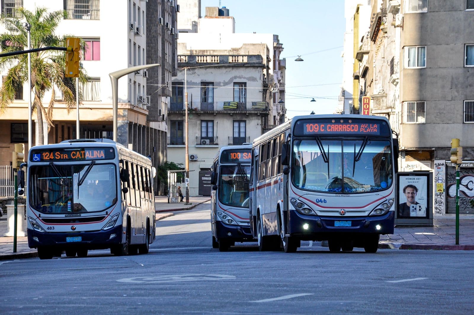 Three city buses with route numbers on their destination signs pulling through a downtown street